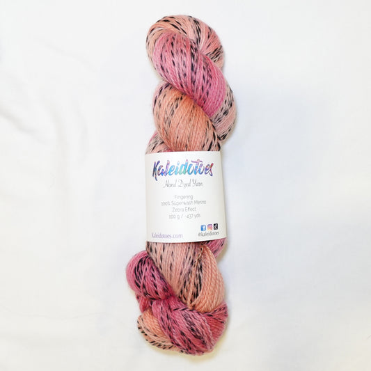 Hand-dyed fingering weight yarn in soft peachy-pink, warm peach, and dusty coral with black twisted through throughout.