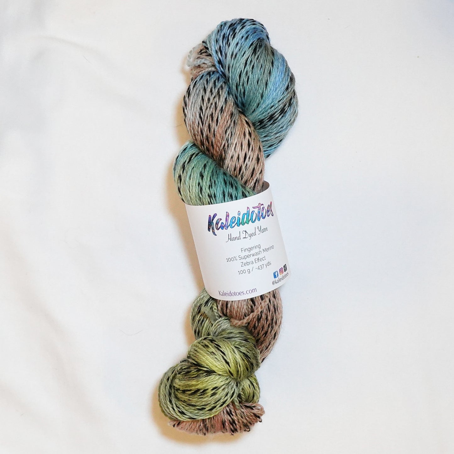 Hand-dyed fingering weight yarn in sand, sage, sky blue, and olive with black threading throughout on a zebra base.