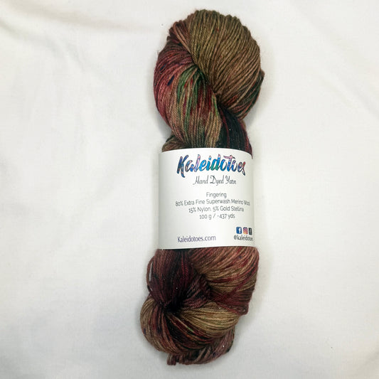 Hand-dyed yarn in warm gingerbread brown, cranberry red, caramel, evergreen, and subtle plum tones with delicate gold metallic flecks.